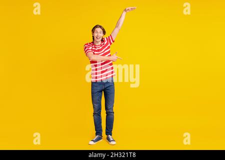 Photo de doux impressionné jeune homme porter un t-shirt rayé mesurer la taille doigt de pointage vide espace isolé couleur jaune fond Banque D'Images