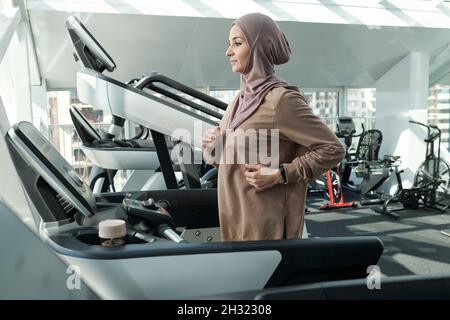 Jeune femme en hijab et en forme d'activewear s'exerçant sur une grande variété d'appareils de sport dans la grande salle de gym moderne Banque D'Images