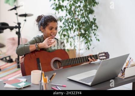 Bonne préadolescence écolière avec guitare en agitant la main à son professeur sur l'écran d'ordinateur portable pendant la leçon de musique en ligne Banque D'Images