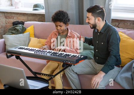 Un jeune professeur de musique se félicitant de son élève jouant un clavier de piano tout en étant assis sur un canapé et en ayant des leçons dans son environnement domestique Banque D'Images