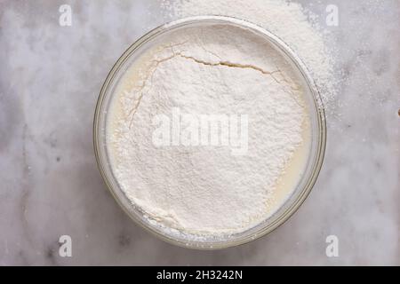 Vue de dessus d'un bol en verre avec de la pâte à frire recouverte de farine tamisée sur une surface en marbre Banque D'Images