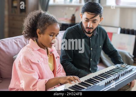 Adorable écolière biraciale touchant les touches du clavier de piano tout en écoutant son professeur de musique pendant la leçon à la maison Banque D'Images