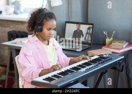 Jolie écolière dans un casque jouant un clavier de piano tout en écoutant les conseils de son professeur pendant la leçon en ligne Banque D'Images