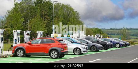 Câble de charge de voiture électrique vers le capot avant rouge d'autres voitures coffre arrière extrémité de hayon connecté à la rangée de points d'alimentation de remplissage baies de stationnement vertes Rugby UK Banque D'Images