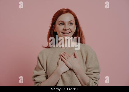 Jeune heureuse femme aux cheveux rouges mettant les mains sur la poitrine tout en se tenant isolée sur fond rose Banque D'Images