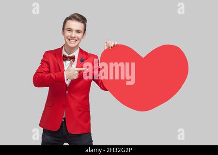 Portrait de l'homme beau et enthousiaste avec une coiffure élégante en rouge tuxedo pointant vers le cœur de grand papier rouge et souriant, exprimant une joie extrême, saluant le jour de la Saint Valentin. studio tourné, fond gris Banque D'Images