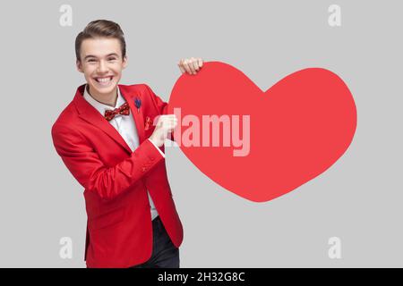 Portrait d'un homme enthousiaste avec une coiffure élégante en smoking rouge et noeud papillon tenant un grand coeur en papier rouge et souriant, exprimant une joie extrême, salutation le jour de la Saint Valentin. photo de studio, fond gris Banque D'Images