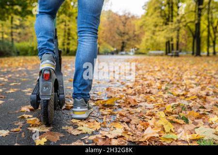 Jambes d'un homme en jeans et baskets debout sur un scooter électrique sur le trottoir de la rue d'automne avec espace de copie.Personne méconnaissable sur un e-scooter de ville moderne.Concept de location de scooters électriques Banque D'Images