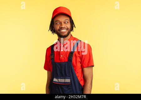 Sourire optimiste constructeur ou chargeur portant l'uniforme, regarder loin avec l'expression heureuse, en attendant de nouvelles commandes, travaille dans l'industrie de service avec plaisir.Studio d'intérieur isolé sur fond jaune Banque D'Images