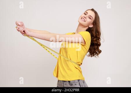 Portrait de profil bonne femme de sourire en dents de souris avec sourire en T-shirt jaune mesurant sa taille avec mètre ruban, satisfait du résultat.Prise de vue en studio isolée sur fond gris. Banque D'Images