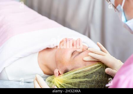 Une esthéticienne utilise des mains gantées pour appliquer une solution chimique de peeling sur le visage du patient. Banque D'Images