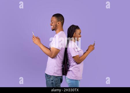Style de vie moderne.Profil vue de côté portrait d'un couple afro-américain positif gai debout dos à dos, tenant et utilisant leur téléphone mobile Banque D'Images