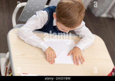 Un adorable jeune garçon de première classe dans un uniforme d'école à la maison tandis qu'isolé à son bureau fait un avion en papier pendant le renfoncement. Mise au point sélective. Portrait Banque D'Images