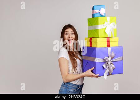 Surprise femme excitée tenant beaucoup de boîtes enveloppées et regardant avec étonnement, choqué par le montage de bonus et de cadeaux, portant T-shirt blanc.Prise de vue en studio isolée sur fond gris. Banque D'Images