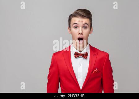 Je n'y crois pas.Portrait de l'homme émotionnel avec une coiffure élégante en smoking rouge et noeud papillon debout avec les grands yeux et la bouche ouverte, regardant l'appareil photo dans un choc étonnement. prise de vue en studio, fond gris Banque D'Images