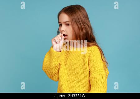 Portrait d'une petite fille malade malsaine ayant des symptômes de grippe, toux, gardant la main près de la bouche, portant un chandail jaune de style décontracté.Studio d'intérieur isolé sur fond bleu. Banque D'Images