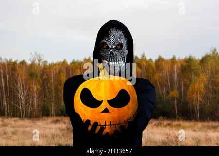 Grim reaper tenant la tête de citrouille d'halloween.Homme dans le masque de la mort avec flamme de feu dans les yeux sur jaune automne nature ensoleillé fond.Conc. Fêtes Halloween Banque D'Images