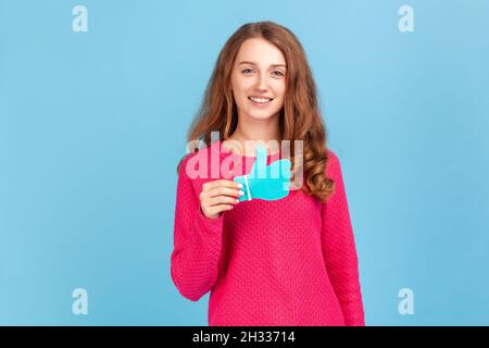 Femme souriante portant un pull-over rose, montrant les pouces vers le haut de l'enseigne de papier bleu, aimer et recommander des postes dans les réseaux sociaux, demandant à évaluer.Studio d'intérieur isolé sur fond bleu. Banque D'Images