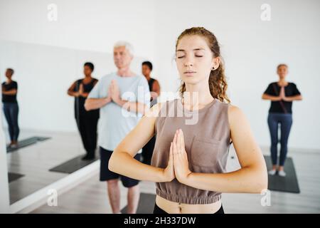 Une jeune femme avec les yeux fermés a joint ses mains méditant en cours de yoga avec d'autres personnes en arrière-plan Banque D'Images
