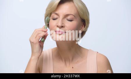 blonde mature femme avec les yeux fermés utilisant le rouleau de jade isolé sur blanc Banque D'Images