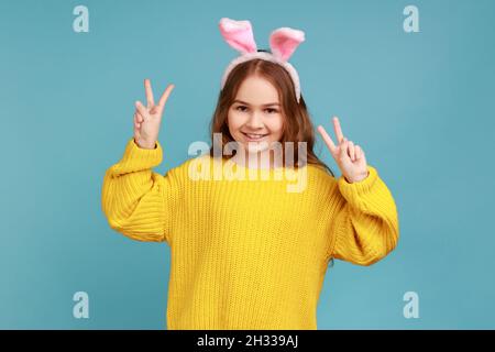 Portrait de petite fille stands avec des oreilles de lapin rose, enfant ayant le plaisir, montrant le signe v à l'appareil photo, portant jaune style décontracté pull.Studio d'intérieur isolé sur fond bleu. Banque D'Images