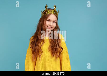 Portrait d'une petite fille mignonne en couronne de diadem doré, regardant l'appareil photo avec un sourire crasseux, portant un chandail jaune de style décontracté.Studio d'intérieur isolé sur fond bleu. Banque D'Images