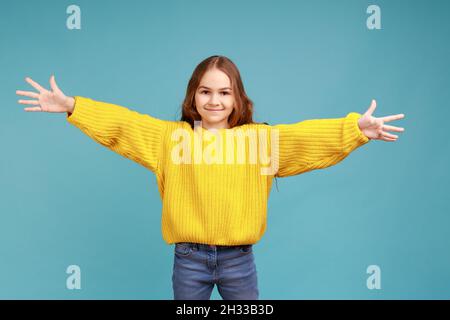 Portrait d'une petite fille adorable et heureuse qui dépasse les mains pour embrasser, donnant des câlins libres et accueillant, en portant un pull jaune décontracté.Studio d'intérieur isolé sur fond bleu. Banque D'Images