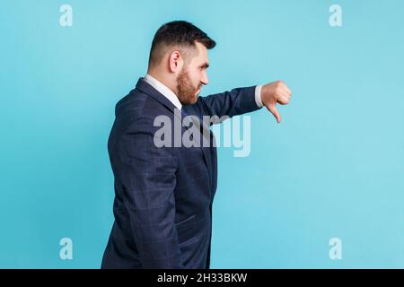 Portrait d'insatisfait beau barbu jeune homme adulte portant un costume officiel montrant les pouces vers le bas, insatisfait du service, mauvais feedback.Studio d'intérieur isolé sur fond bleu. Banque D'Images