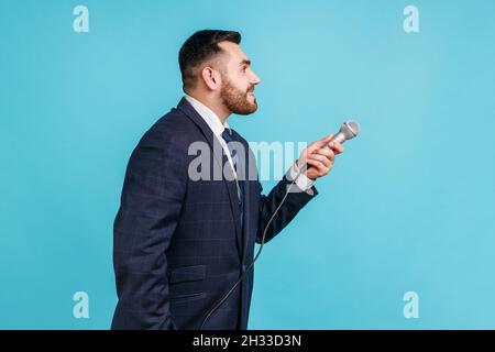 Portrait de la vue latérale d'un jeune adulte barbu portant un costume officiel debout et offrant un microphone, posant des questions, des entretiens.Studio d'intérieur isolé sur fond bleu. Banque D'Images