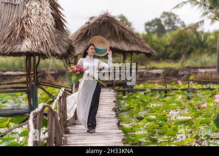 Portrait de la belle femme vietnamienne avec chapeau traditionnel vietnam tenant le lotus rose marchant sur le pont en bois dans le grand lac de lotus, vietnam, ai Banque D'Images