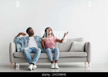 Portrait d'un couple afro-américain souffrant de la chaleur estivale, femme sueur tenant la télécommande allumer la climatisation, famille assis sur co Banque D'Images