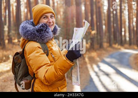 Jeune femme souriante dans une veste chaude orange.Une femme d'origine ethnique caucasienne attrayante marche dans le parc avec un sac à dos.Elle regarde la carte.Randonnée en t Banque D'Images