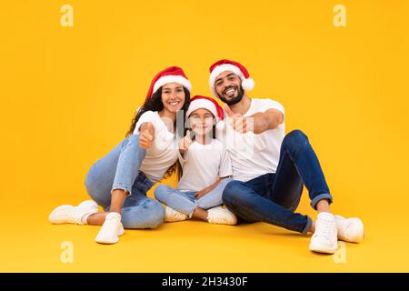 Bonne famille arabe à Santa Hats posant montrant des Thumbs Up geste approuvant les traditions de Noël assis sur le fond jaune de studio.Nous aimons Noël Banque D'Images