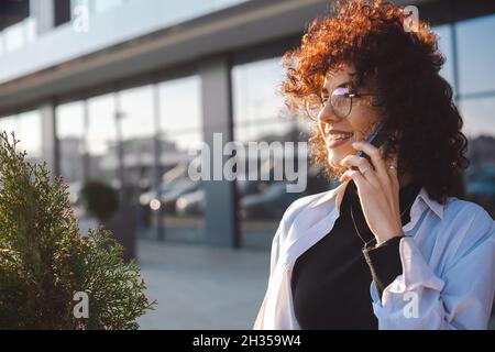 Portrait d'une femme d'affaires ayant des conversations téléphoniques.Femme aux cheveux bouclés et aux lunettes souriant en toute confiance à la ville.Communication mobile. Banque D'Images