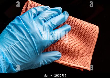 Une main dans un gant de protection bleu essuyant la poussière, la saleté d'une surface noire avec un chiffon, désinfecter la surface, gros plan.Concept de nettoyage de maison.Disin Banque D'Images
