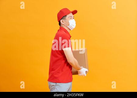 Livreman employé en casquette rouge t-shirt blanc uniforme masque gants tenir boîte en carton isolée sur fond jaune Banque D'Images