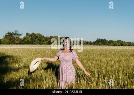 Photo de style de vie d'une belle femme aux cheveux longs, tenant un chapeau de soleil, marchant dans le champ de blé ou d'orge Banque D'Images