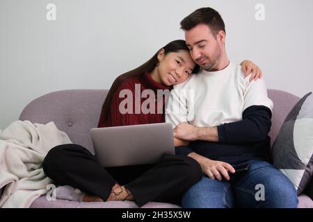 Un jeune couple se détend sur un canapé à la maison à l'aide d'un ordinateur portable et embrassant. Banque D'Images