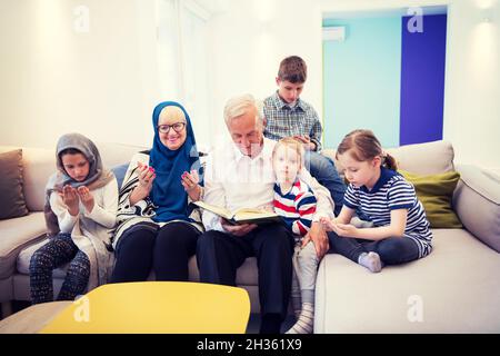 La famille musulmane moderne des grands-parents avec leurs petits-enfants la lecture de Coran et priant ensemble sur le canapé avant de dîner iftar ramadan lors d'une fête à la maison Banque D'Images