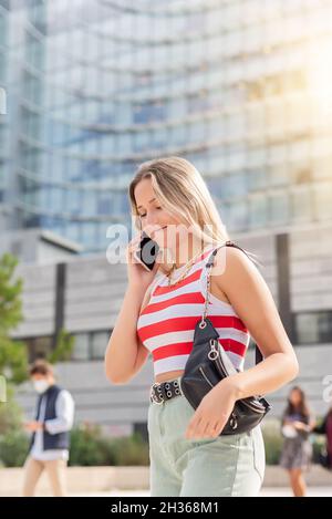 Jeune femme blonde avec sac à main marchant dans la rue et parlant au téléphone Banque D'Images