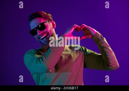 Photo portrait de l'homme souriant portant un verre de soleil montrant le coeur signe avec les doigts sur la date isolée sur fond violet foncé Banque D'Images