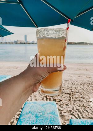 Femelle méconnaissable tenant un verre à cocktail à la main contre un fond marin, prise de vue courte d'une personne allongé sur un lit de soleil avec une boisson rafraîchissante froide et du rel Banque D'Images