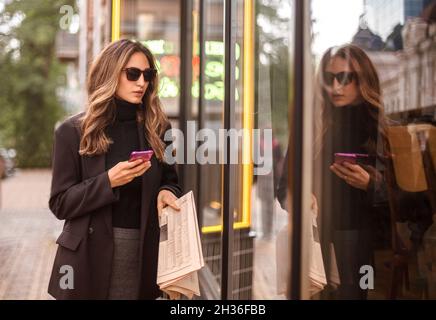 Jeune femme élégante et tendance portant des lunettes noires marchant dans la rue de la ville et regardant dans la fenêtre du magasin, femme d'affaires confiante avec newspa Banque D'Images