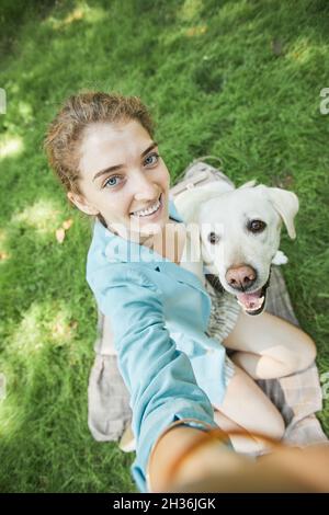 Portrait vertical en grand angle d'une jeune femme souriante prenant le selfie avec son chien à l'extérieur tout en appréciant la marche dans le parc ensemble Banque D'Images