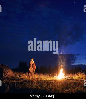 Ciel nuageux de nuit sur une colline herbeuse avec tentes de camp, feu de camp et randonneurs.Jeune homme et femme touristes appréciant la vue du ciel bleu de nuit.Concept de relation, de camping de nuit et de voyage. Banque D'Images