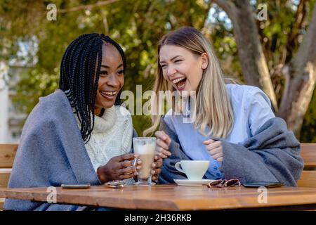 Deux jeunes amies qui apprécient le café sur la terrasse du café, qui s'assoit à une table, bavardent et rient à voix haute.Diversité. Banque D'Images