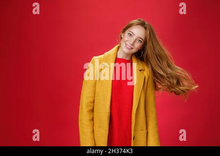 Charmante femme à tête rouge avec des taches de rousseur qui agite les cheveux en souriant large tête inclinable comme une coupe de cheveux volante dans l'air en posant ravie et insouciante sur le rouge Banque D'Images