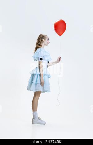 Vue latérale d'une jeune fille portant une robe d'Halloween, costume de personnage de film debout avec ballon rouge à la main isolé sur fond bleu Banque D'Images