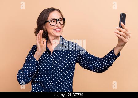 Photo de bossy impressionné vieille dame appel vidéo téléphone porter des lunettes chemise bleue isolée sur fond beige couleur Banque D'Images