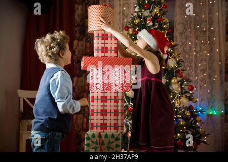 Les enfants adorés et heureux sourient, jouant avec une pile de cadeaux de Noël.Salon joliment décoré avec des lumières et un sapin de Noël. Banque D'Images